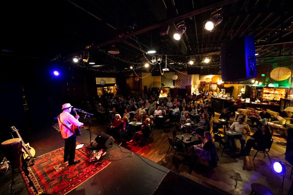 A person wearing a hat, white longsleeve shirt and black pants performs to a seated crowd in a large venue. A person wearing a hat, white longsleeve shirt and black pants performs to a seated crowd in a large venue.