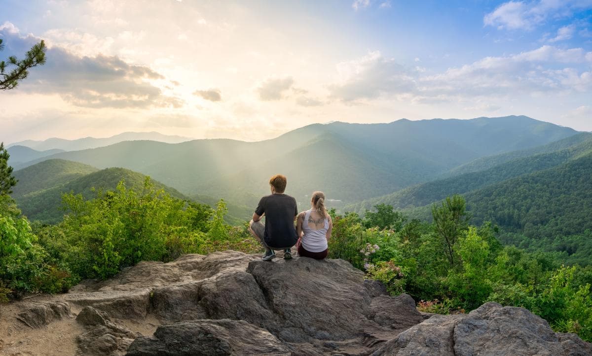 Two people sit on a rock overlooking a mountain range in the foreground with another mountain range and setting sun in the background. Two people sit on a rock overlooking a mountain range in the foreground with another mountain range and setting sun in the background.